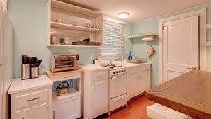 Cocina compacta con electrodomésticos blancos, incluyendo estufa, horno y refrigerador. Hay repisas sobre la estufa con vajilla y una pequeña encimera con tostadora y utensilios.