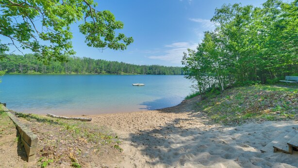 A sandy beach with a wooden bench and a small boat on calm water. - Indoor Pool Access: Cabin in Lakes of the North! (Elmira)