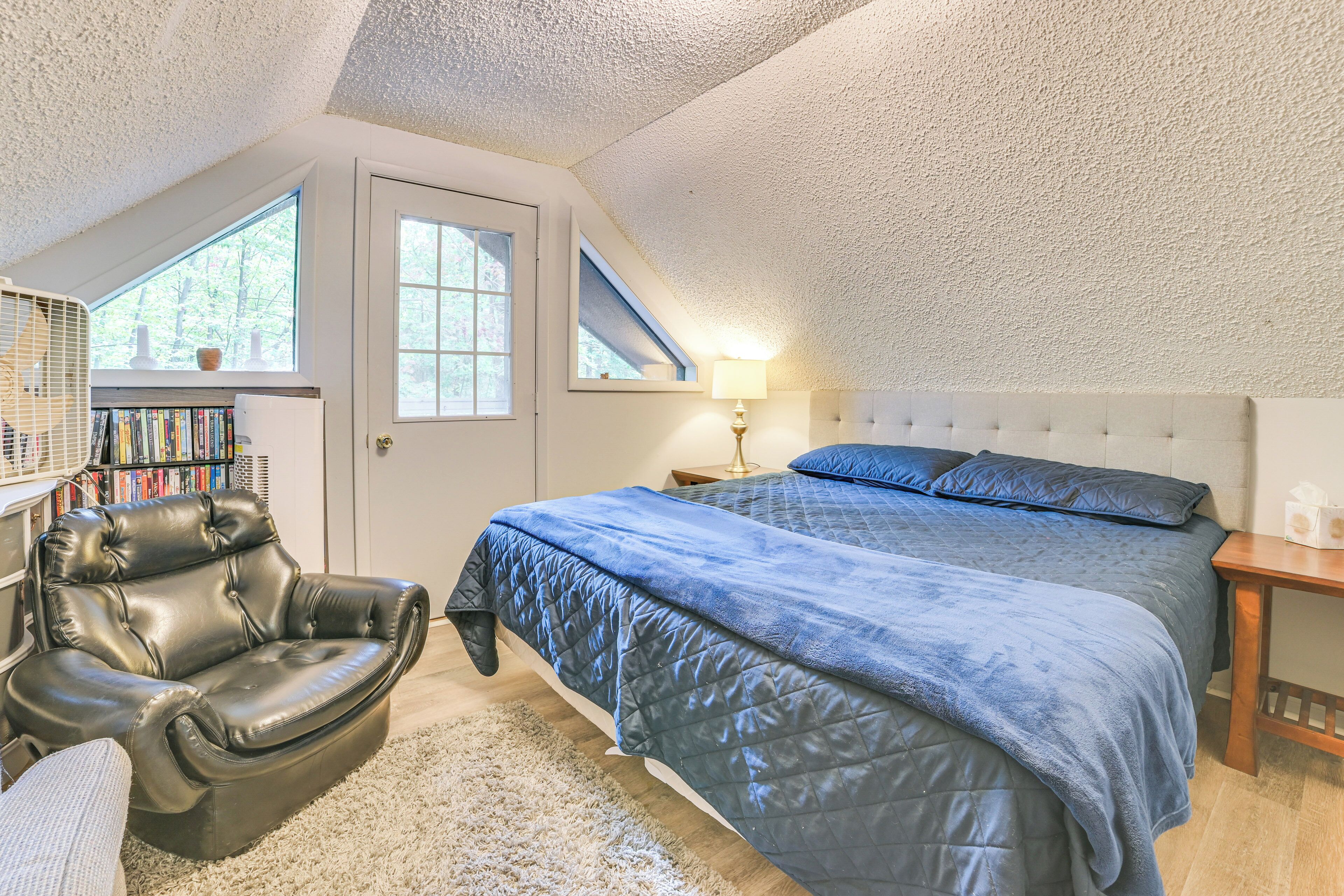 A bedroom with a large bed, a leather chair, a bookshelf, and a window with a view of trees.