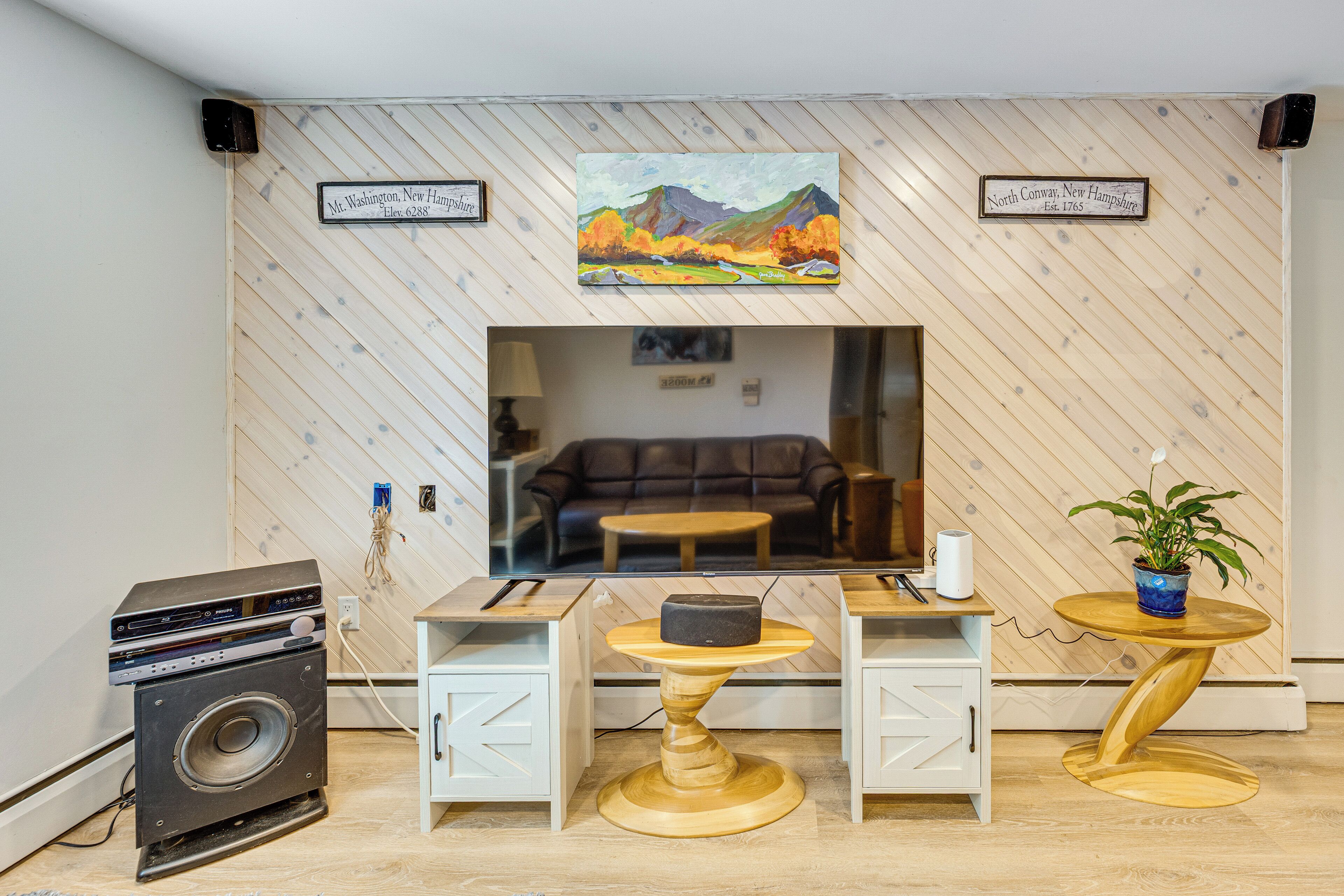 A room with a wooden herringbone wall, a painting of a landscape, a sofa, a coffee table, a small table with a speaker, and a washing machine.
