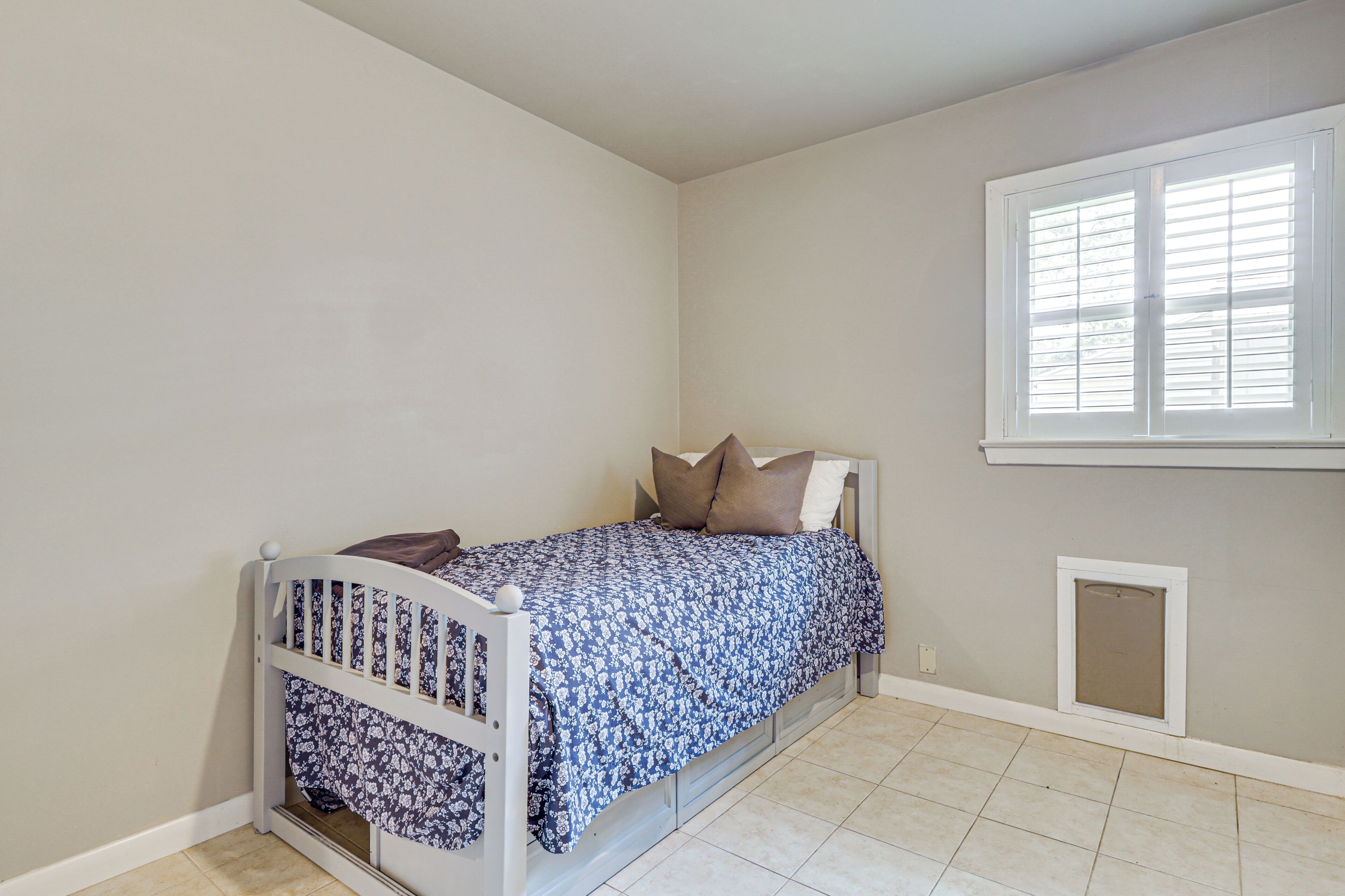 A bedroom with a single bed, a window with blinds, a small vent, and a tiled floor.