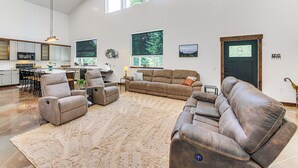 A spacious living room with a large sectional sofa, a recliner, and a coffee table. There is a kitchen in the background with a dining area. - Relax, Reconnect & Explore: Modern Idaho House! (Naples)