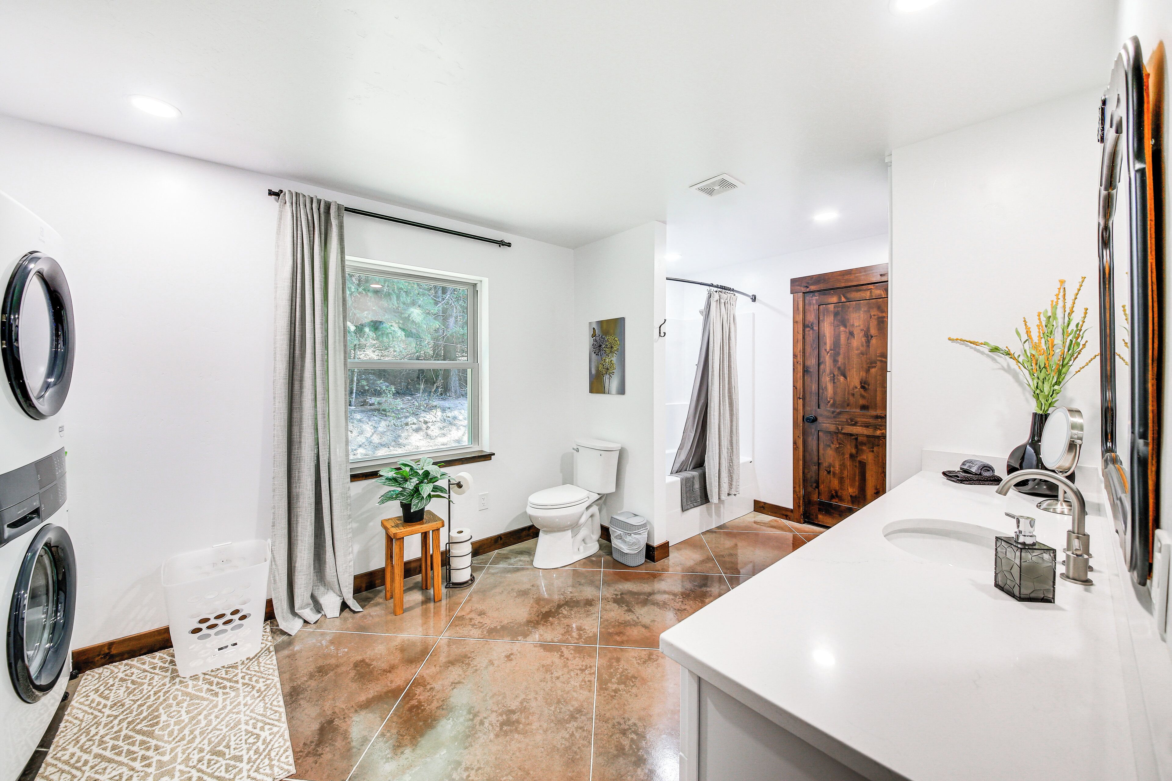 A modern bathroom with a large window, a built-in washing machine, a sink with a mirror, and a shower area.