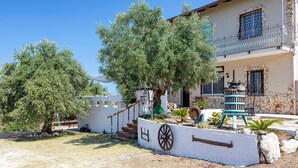 Une maison à deux étages avec une façade en pierre, un balcon orné d’une balustrade en fer forgé et un petit jardin clos par une clôture en bois, où se trouve une brouette.