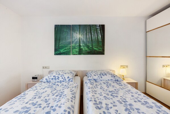 A room with two beds, a wall-mounted painting of a forest, bedside tables with lamps, and a small bedside clock.