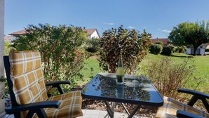 Een terras buiten met een glazen tafel, twee stoelen met geruite kussens, en een fles wijn.