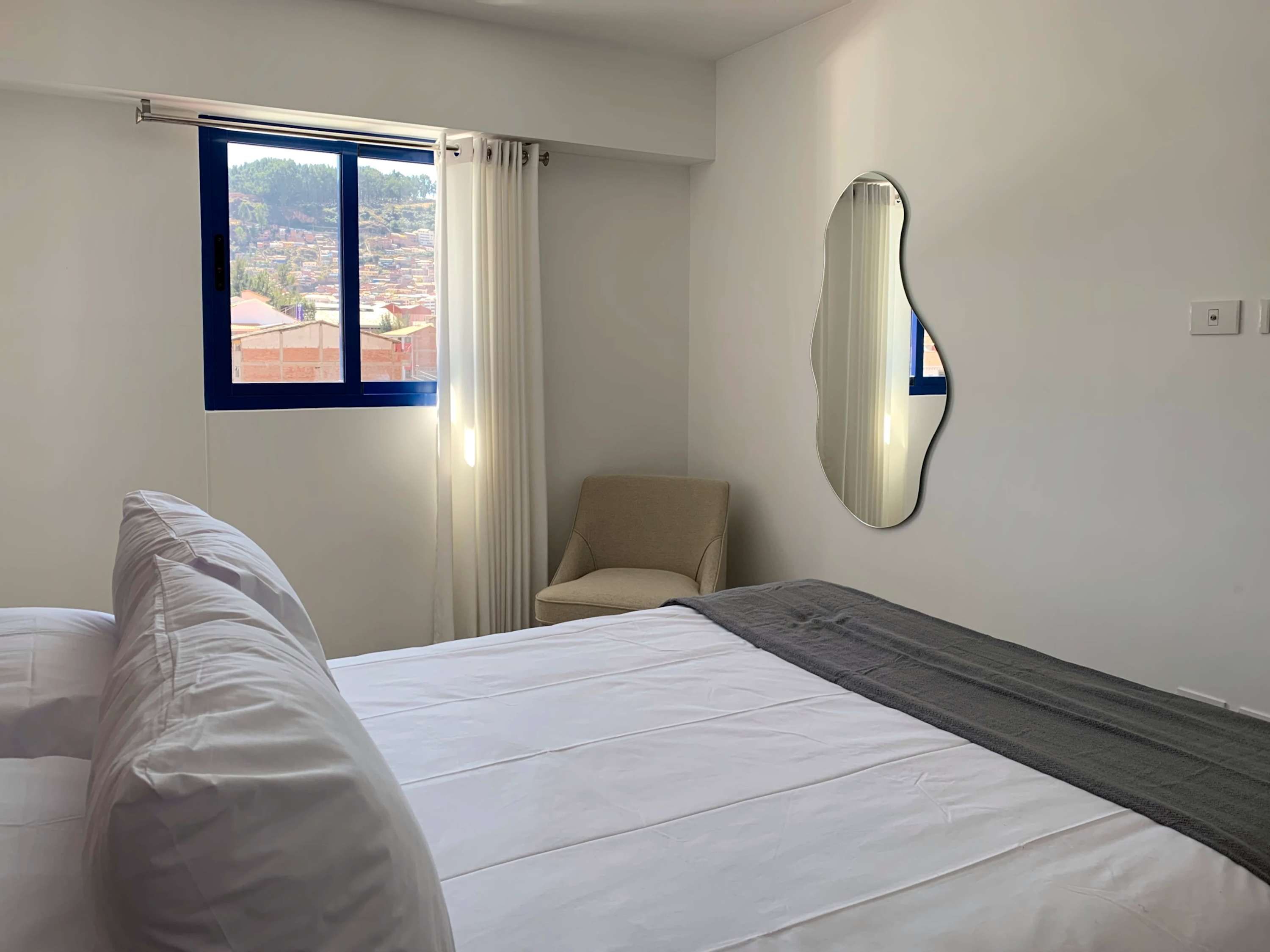 A hotel room with a bed, a chair, a window with curtains, and a wall-mounted mirror.