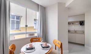 En matplats med ett bord dukat för två personer, ett kök med moderna vitvaror och utsikt över omgivningen genom ett stort fönster.