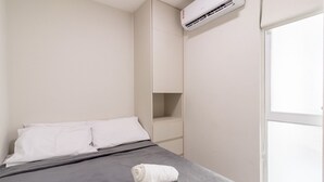 Una habitación de hotel minimalista con una cama, una almohada blanca, una toalla blanca enrollada y un equipo de aire acondicionado integrado.