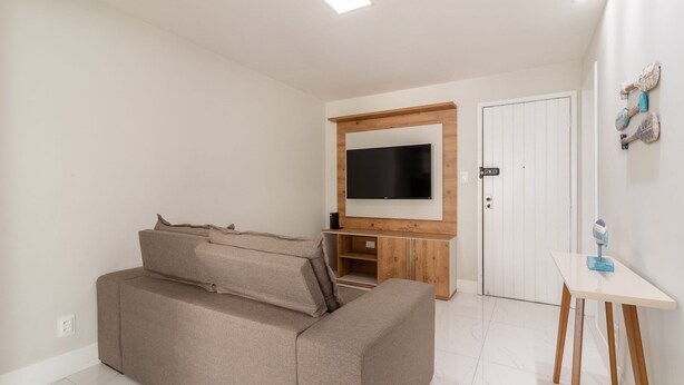 A modern living room with a beige sofa, a flat-screen TV mounted on a wooden wall, a small table with decorative items, and a white door. - Beautiful Apart in Barra 110m From Beach Csf0202 (Salvador)