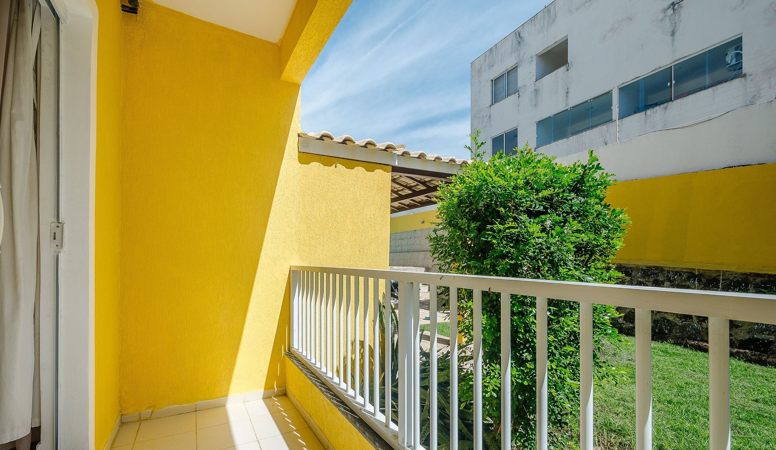 A yellow exterior wall with a white balcony railing, a small tree, and a building with large windows in the background.