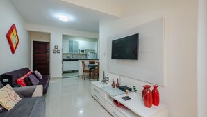 Un salon moderne avec un canapé, un téléviseur et un meuble de rangement pour les appareils électroniques. On aperçoit une cuisine au fond.
