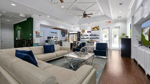 A spacious living area with a beige sofa, a coffee table, and a ceiling fan. There is a kitchen area with a refrigerator and a sign that says "Padoka."