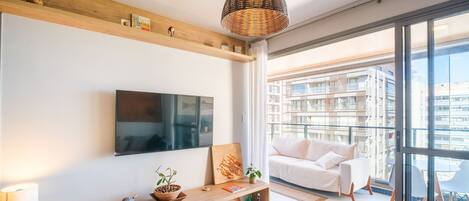 A modern living room with a flat-screen TV, a sofa, a wooden console table with decorative items, and a large window with a city view.