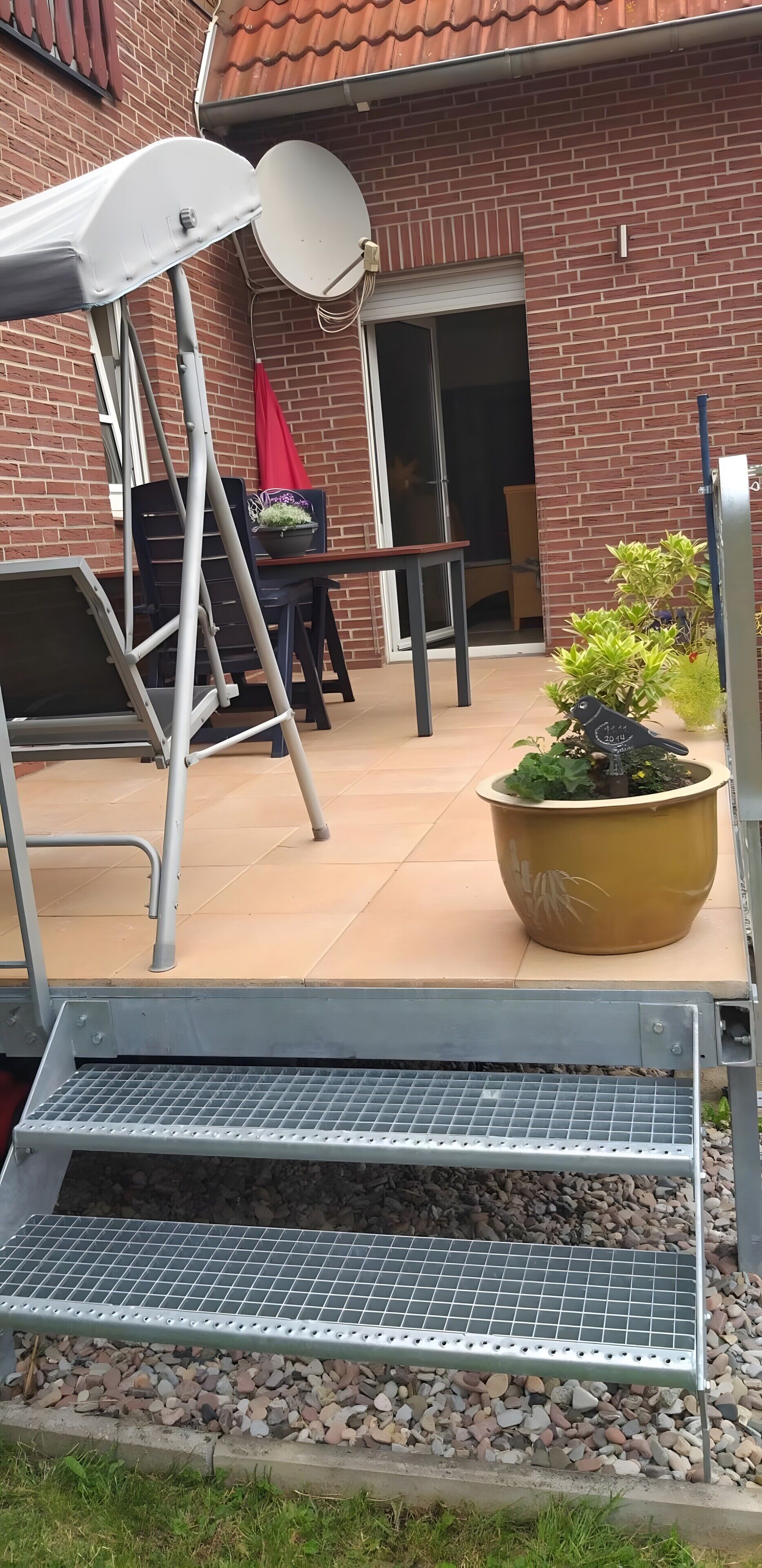 Eine Terrasse mit einer Metallbank, einem Blumentopf, einer Satellitenschüssel und einem Backsteinhaus im Hintergrund.