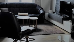 Een moderne woonkamer met een hoekbank, een zwarte bureaustoel, twee kleine ronde tafels, een flatscreen televisie en een accentmuur met een stenen textuur.