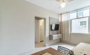 Una sala de estar moderna con un televisor de pantalla plana montado en la pared, una mesa de comedor con sillas y vistas al exterior a través de una ventana con cortinas.