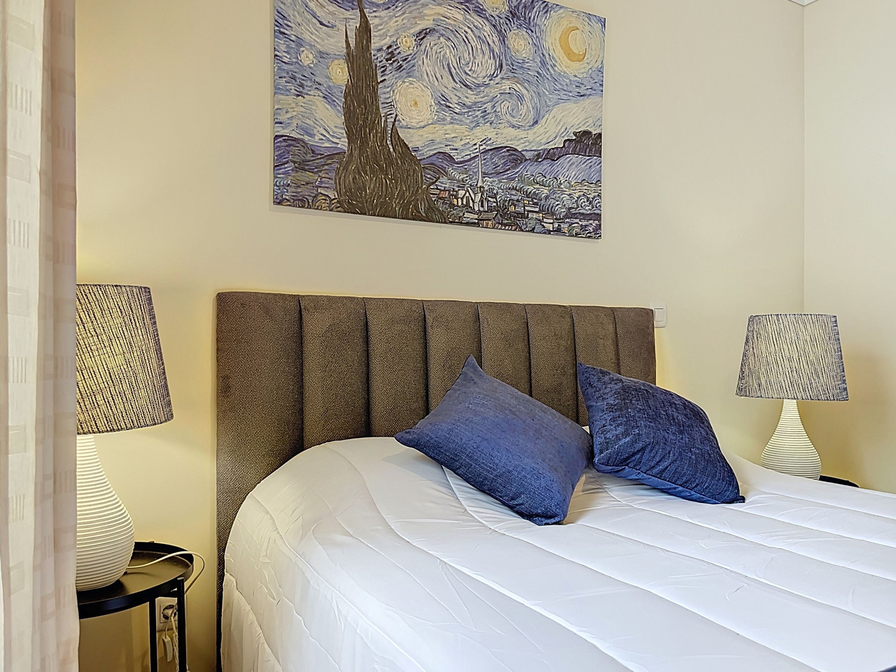 A neatly made bed with a white duvet and two blue pillows, a brown upholstered headboard, and a painting of a night sky above it.