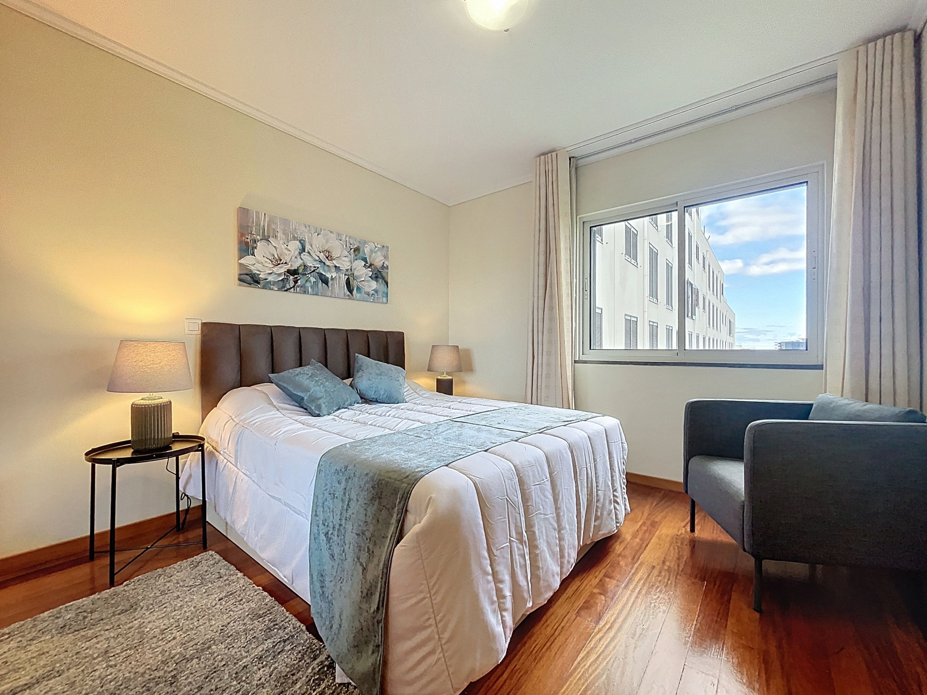 Une chambre d’hôtel moderne avec un grand lit, deux tables de chevet, une chaise, une fenêtre donnant sur la ville et un tableau au mur.