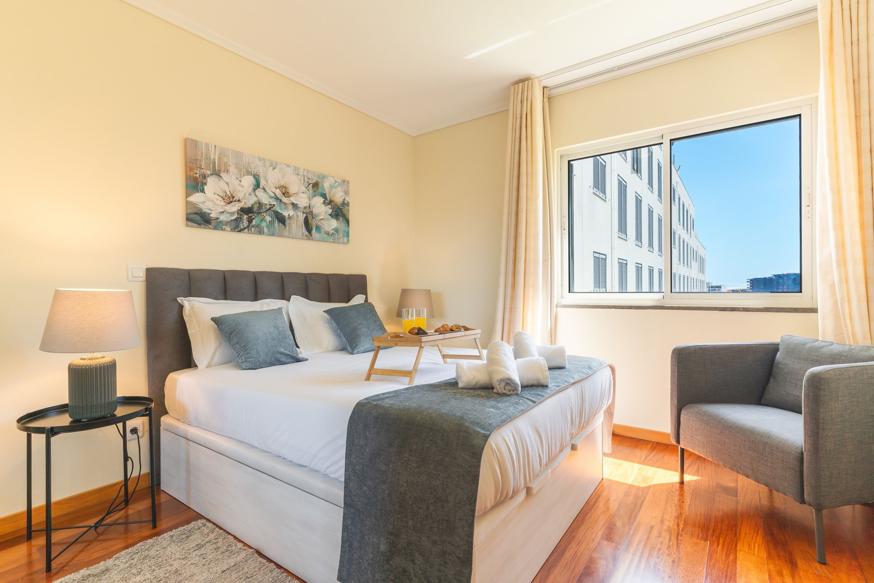 A hotel room with a bed, bedside table, lamp, chair, and a large window with a view of buildings.