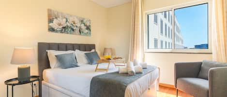 A hotel room with a bed, bedside table, lamp, chair, and a large window with a view of buildings.
