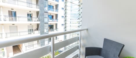 A balcony with a wicker chair, a small table with plates and glasses, and a view of a multi-story building.