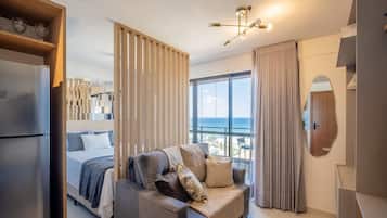 Quarto de hotel moderno com cama, sofá, janela grande com vista e luminária no teto.