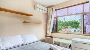 A bedroom with a bed, a desk, a chair, a window with a view of buildings, and an air conditioning unit. - Modern Apartment 1km From Ondina Beach Slo0301 (Salvador)