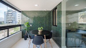 Un balcon avec une table ronde, quatre chaises, un pot de fleurs et une vue sur le paysage urbain, visible à travers de grandes fenêtres.