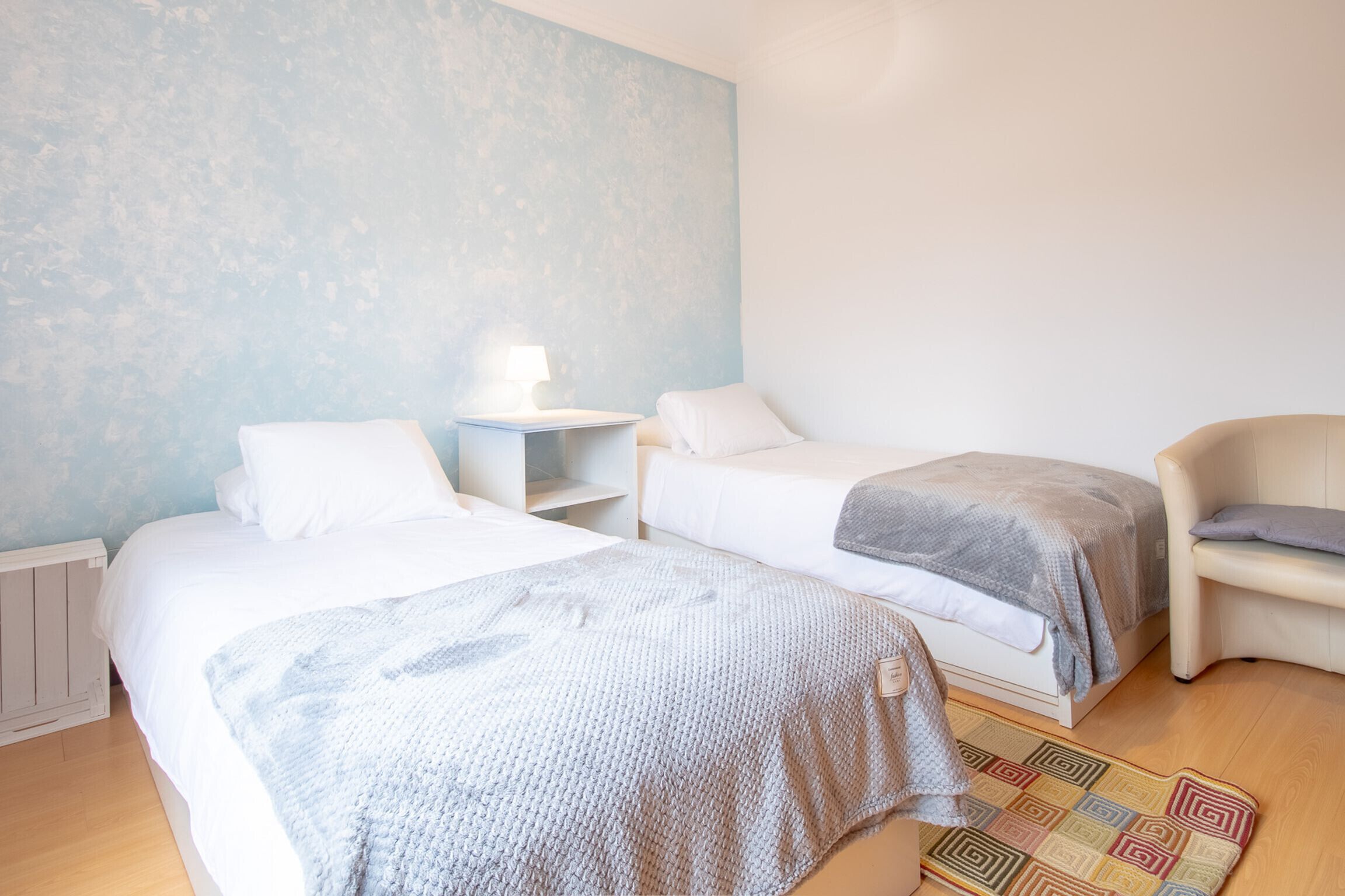 A hotel room with two beds, a bedside table with a lamp, a chair, and a patterned rug.