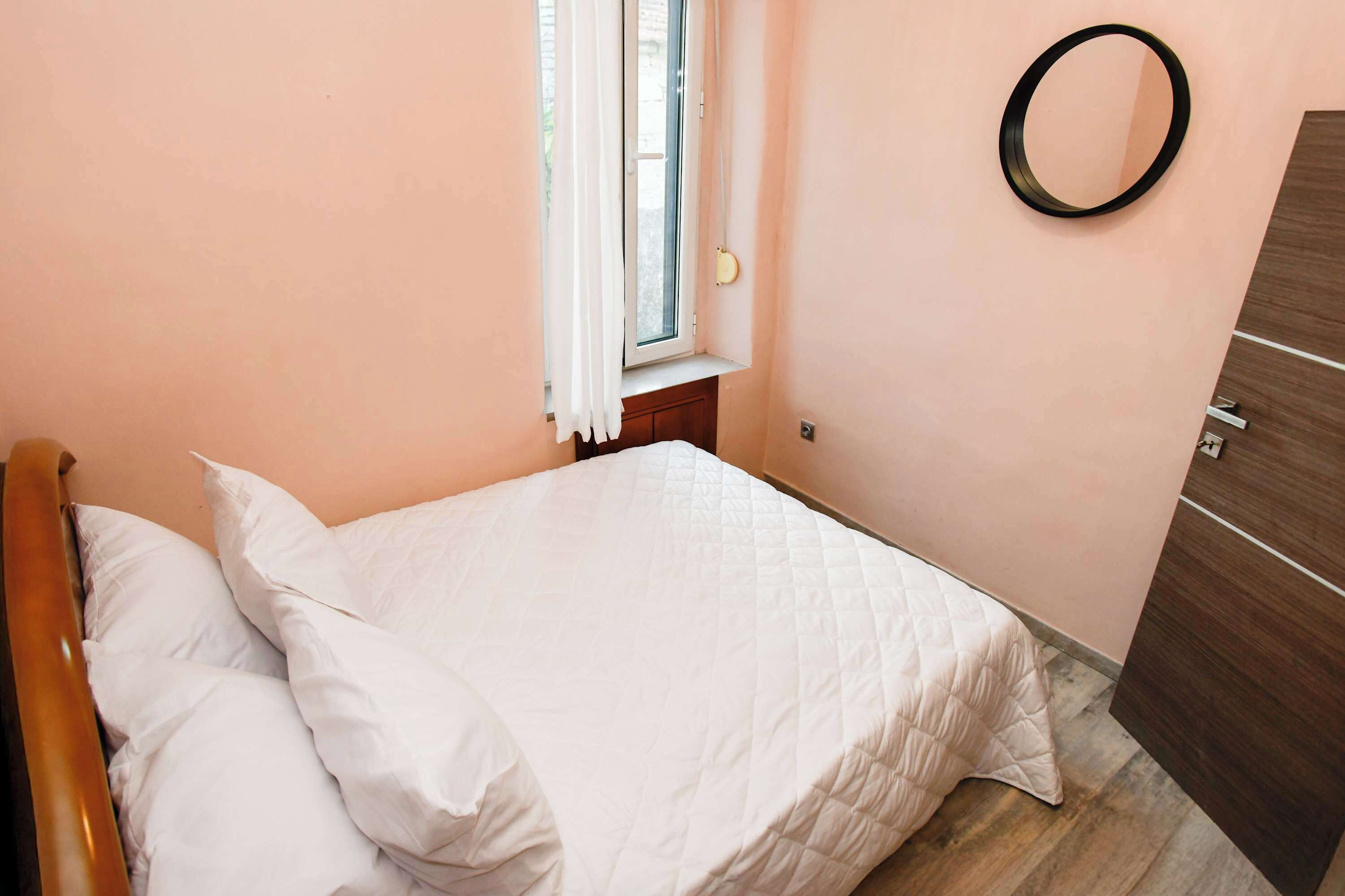 Una cama bien hecha con ropa de cama blanca en una habitación con paredes color melocotón, suelo de madera y una ventana pequeña con cortinas blancas.