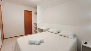 A bedroom with a bed, two pillows, a nightstand, a wooden door, and a white wall. - Nui Supreme Beach Living  (Ipojuca)