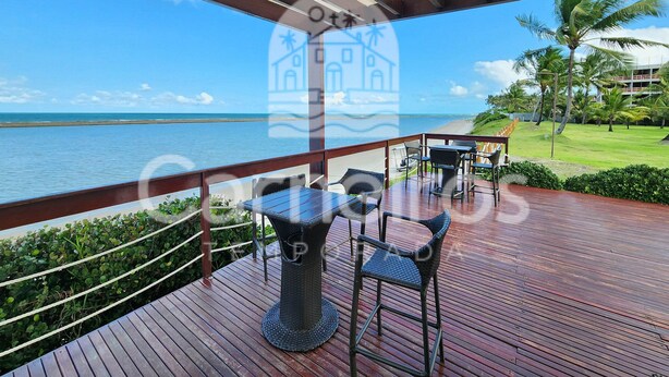 A wooden deck with a view of the ocean, a palm tree, and a building in the background. - Nui Supreme Beach Living  (Ipojuca)
