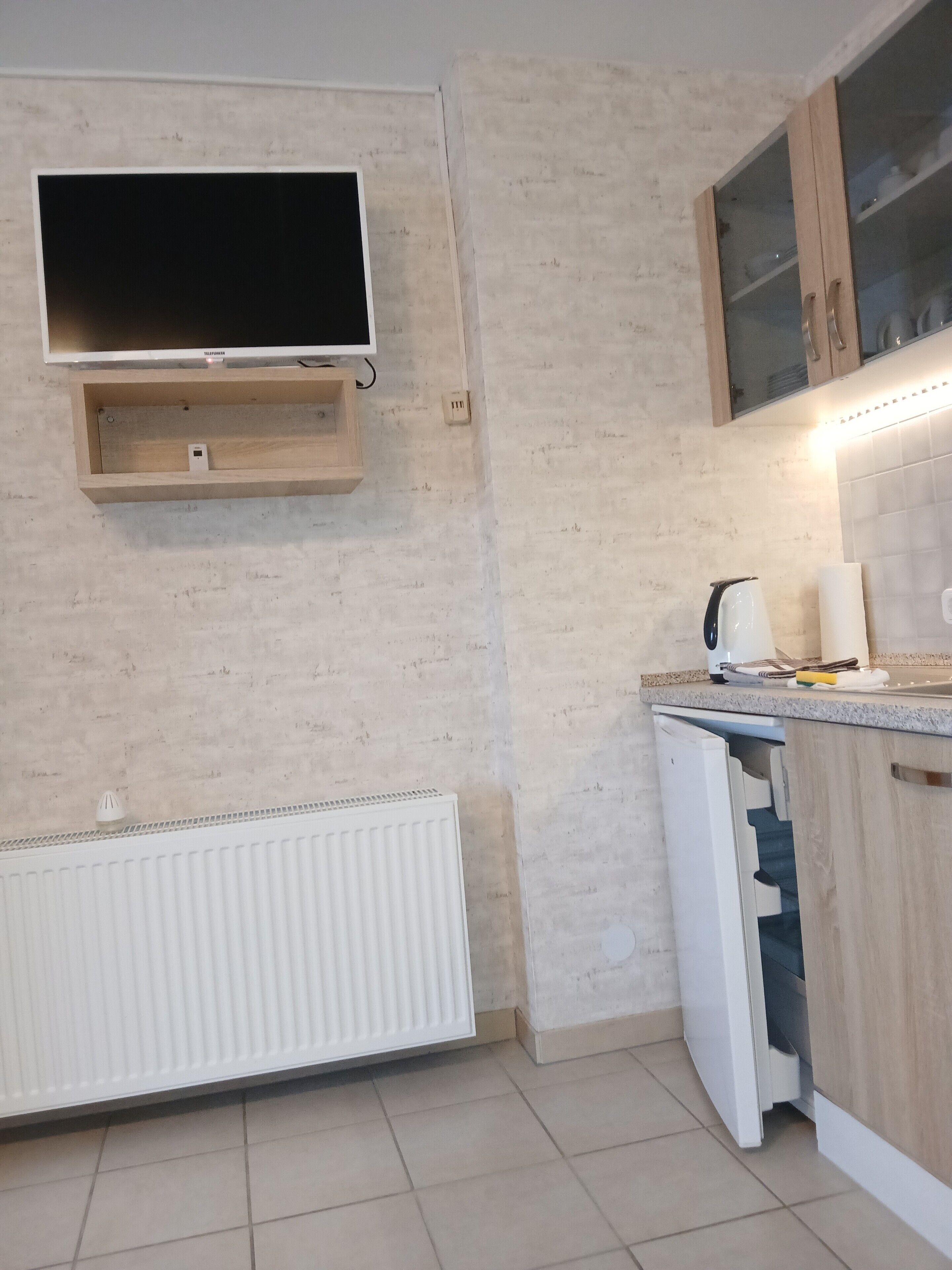 A kitchen with a built-in TV, wooden cabinets, a kettle, and a white refrigerator.