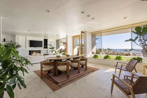 A spacious living area with a dining table, comfortable seating, a flat-screen TV, and large windows offering a view of the outdoors. - Casa Azul by Oraklus (Malibu)