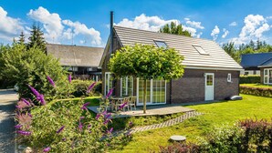 Une maison moderne avec un toit en pente, une véranda en verre et un jardin bien entretenu agrémenté de fleurs violettes.
