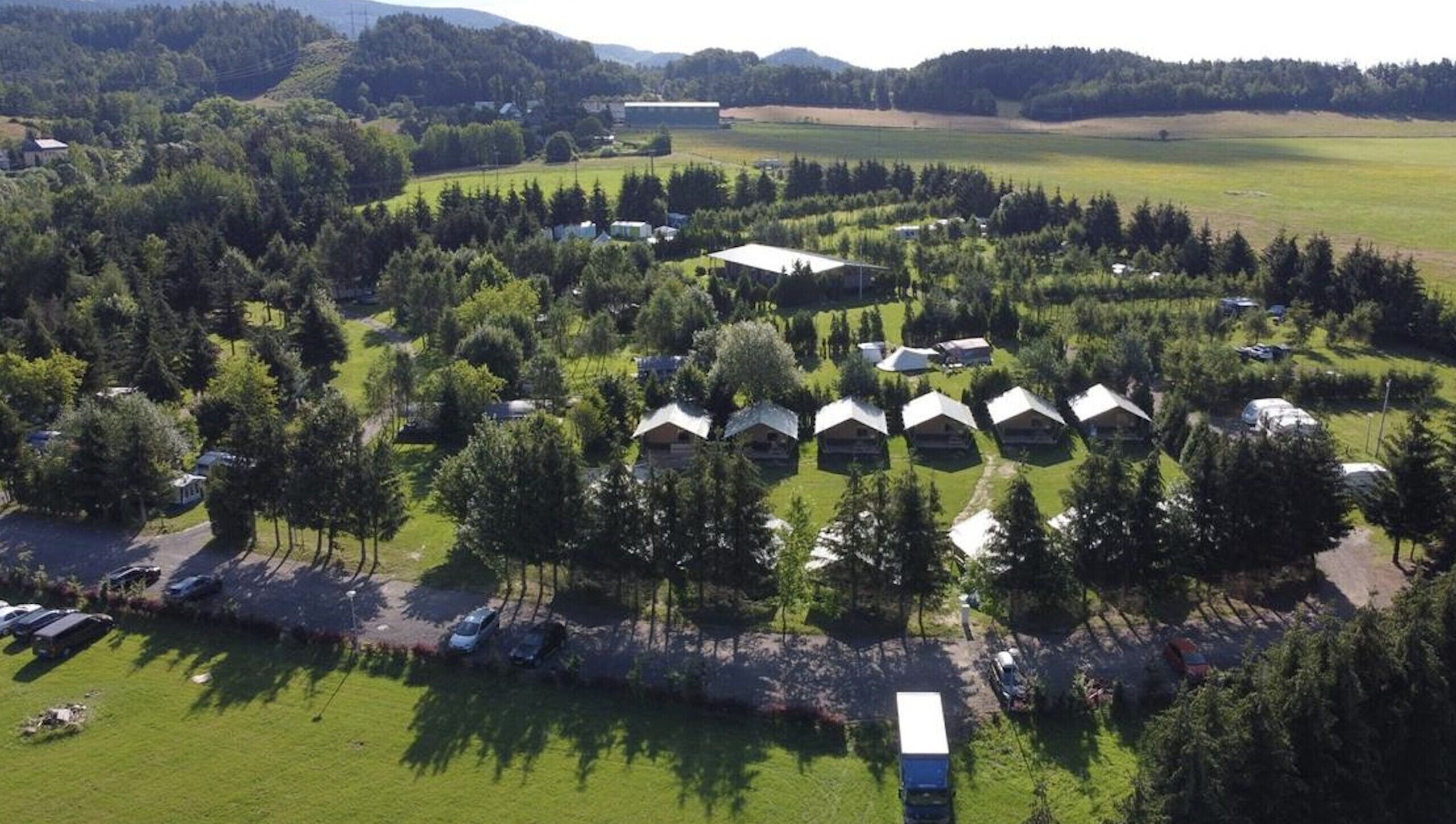 Une vue aérienne d’un terrain de camping avec plusieurs cabanes disposées en demi-cercle, entourées d’une forêt dense et de vastes champs ouverts.