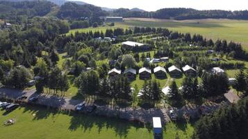 Une vue aérienne d’un terrain de camping avec plusieurs cabanes disposées en demi-cercle, entourées d’une forêt dense et de vastes champs ouverts.