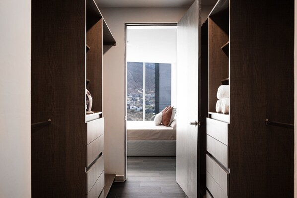 A narrow hallway leading to a bedroom with a large window, wooden wardrobe, and a bed with pillows.