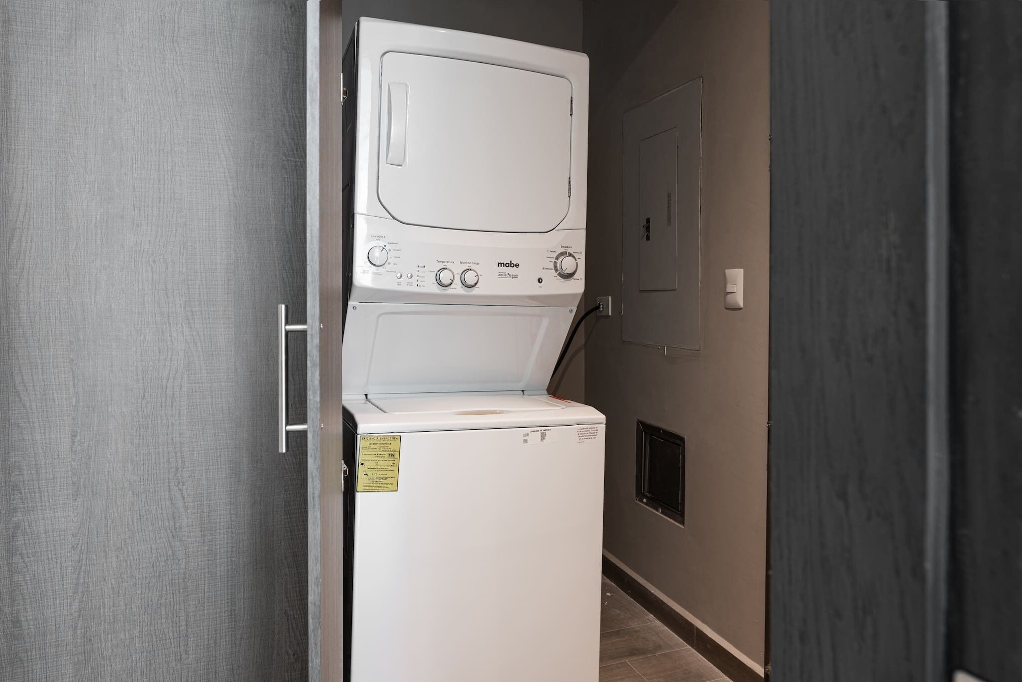 A laundry room with a stacked washer and dryer, a door, and a wall-mounted electrical outlet.
