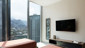 Una habitación de hotel moderna con un amplio ventanal que ofrece vistas de la ciudad, un televisor de pantalla plana montado en la pared y una mesa consola de madera con un control remoto.