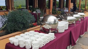 Un buffet est dressé avec des tasses et des assiettes blanches, ainsi qu’un grand pot en argent, le tout placé sur une table recouverte d’une nappe bordeaux.