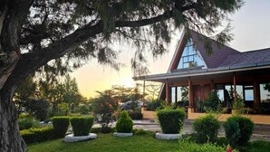 Une maison moderne avec un toit en pente, de grandes fenêtres et un jardin bien entretenu agrémenté de haies et d’un arbre.