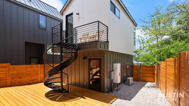Una casa moderna de dos pisos amb una escala espirall que condueix a un balcó, una terrassa de fusta i un pati tancat per una tanca.