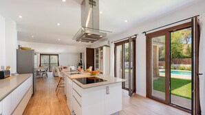A modern kitchen with a central island, a dining area with a table and chairs, and a view of a backyard with a pool. - VILLA SON TONI (Crestatx)