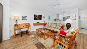 A living room with a sofa, coffee table, and a wooden side table. There is a ceiling fan, a kitchen area with cabinets, and a dining table with chairs. - Big Island Kona Westwind 12 2 Bedroom Condo (Kailua-Kona)