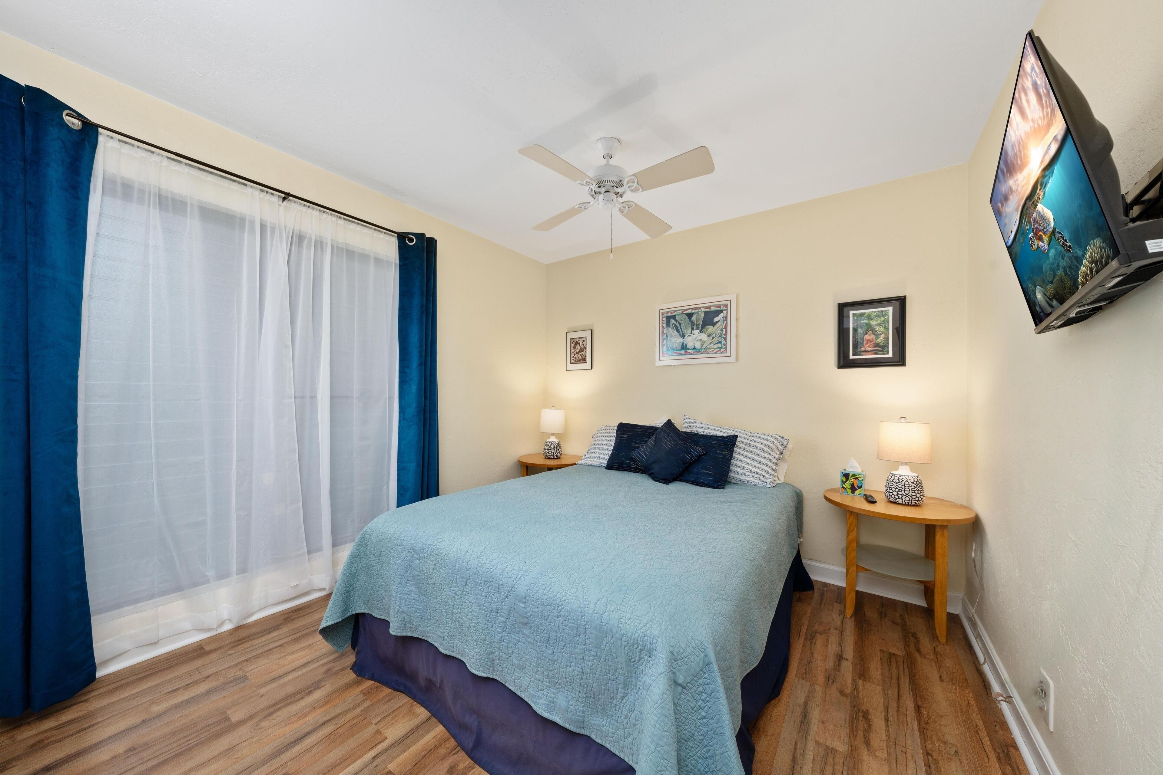 A bedroom with a bed, a ceiling fan, a TV, and a window with curtains.