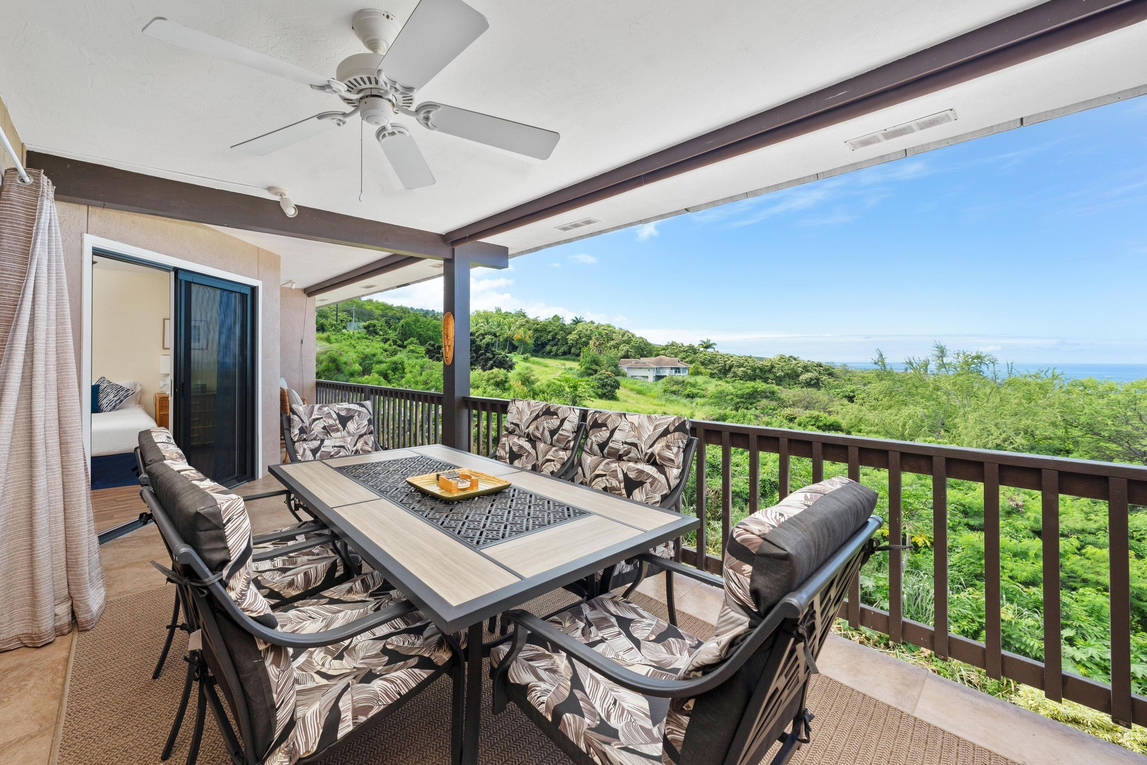 A balcony with a table and chairs, a ceiling fan, and a view of greenery and a house in the distance.