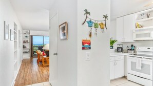 A modern kitchen with white cabinets, a built-in oven, and a microwave. There is a dining area with a table and chairs, and a view of the ocean through a large window. - Big Island Kona Westwind 12 2 Bedroom Condo (Kailua-Kona)