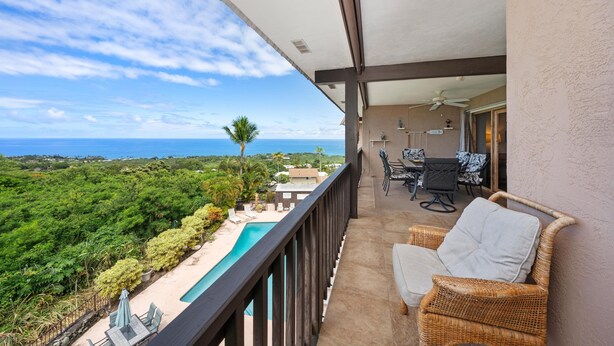 A balcony with a view of a swimming pool and ocean. There is a table and chairs on the balcony. - Big Island Kona Westwind 12 2 Bedroom Condo (Kailua-Kona)
